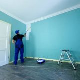 Man in a working overall is painting the wall in white color